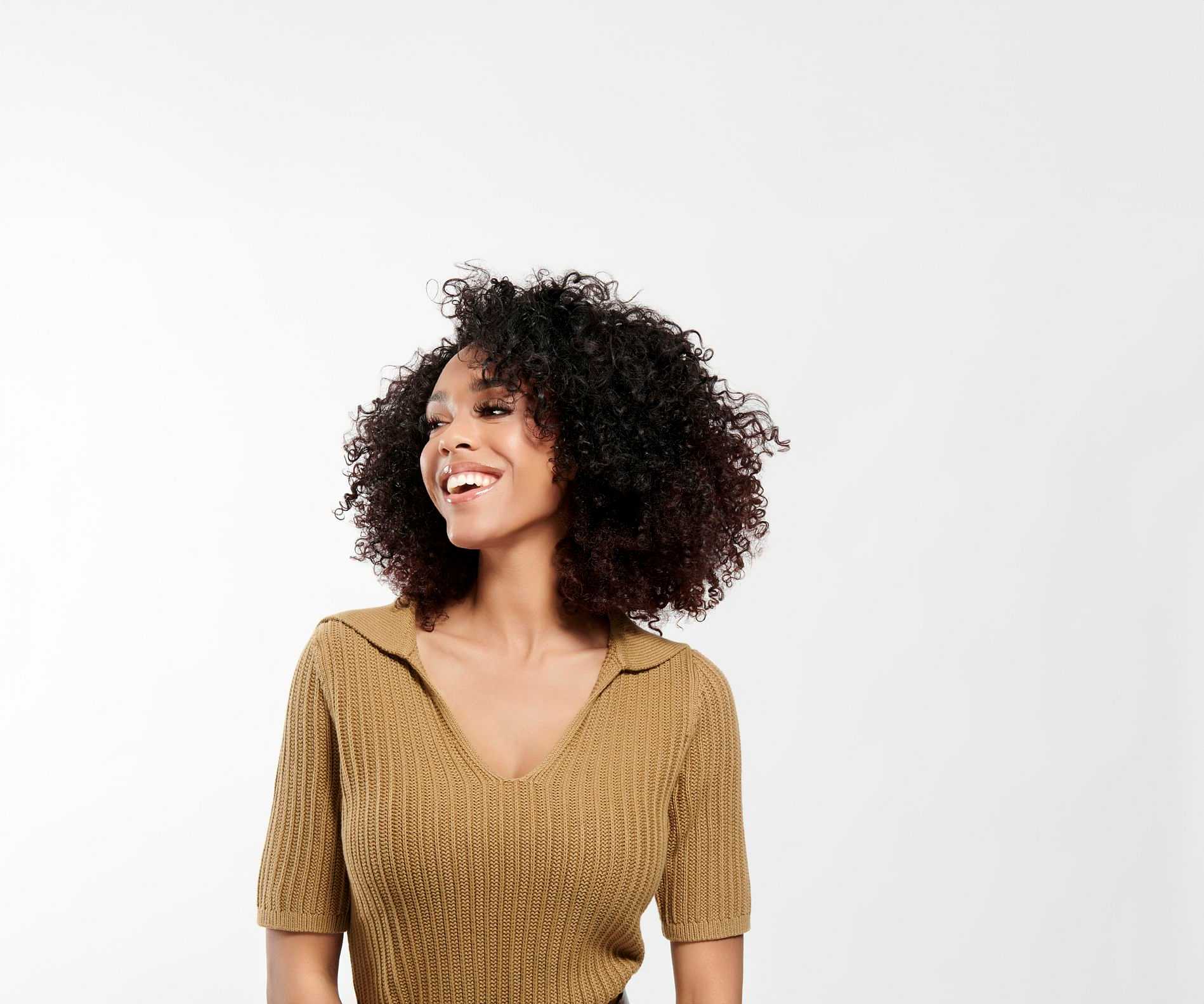 Smiling woman with curly hair wearing a brown sweater against a plain background.