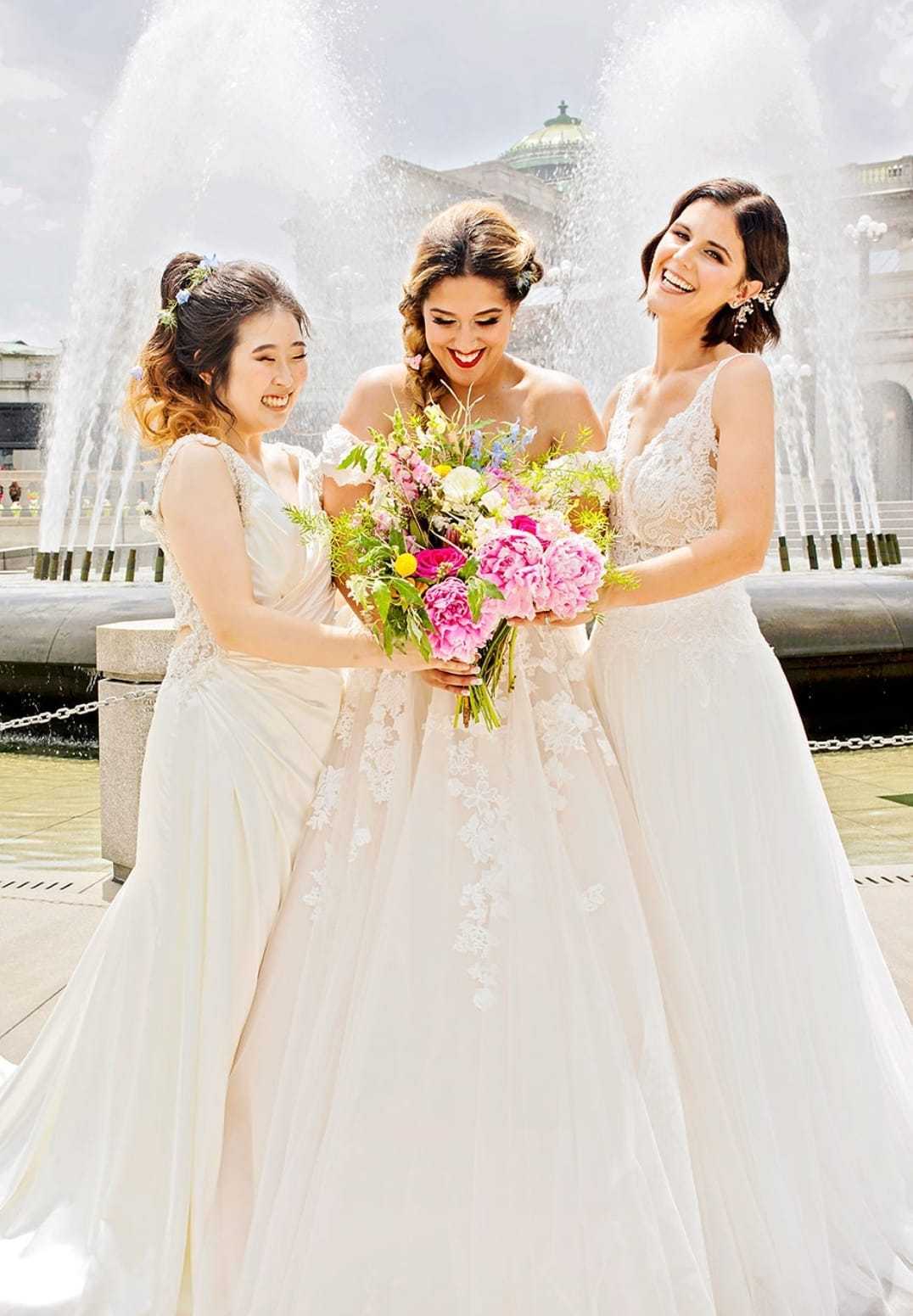 Bridesmaids joyfully admire bride's vibrant bouquet in front of a large fountain.