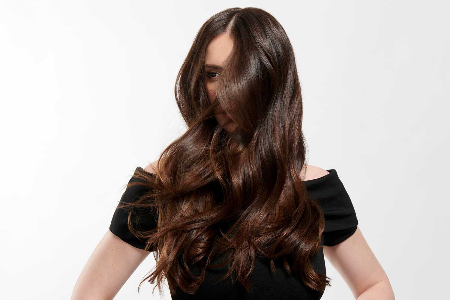 Woman with long, flowing dark hair covers her face, standing against a plain background.