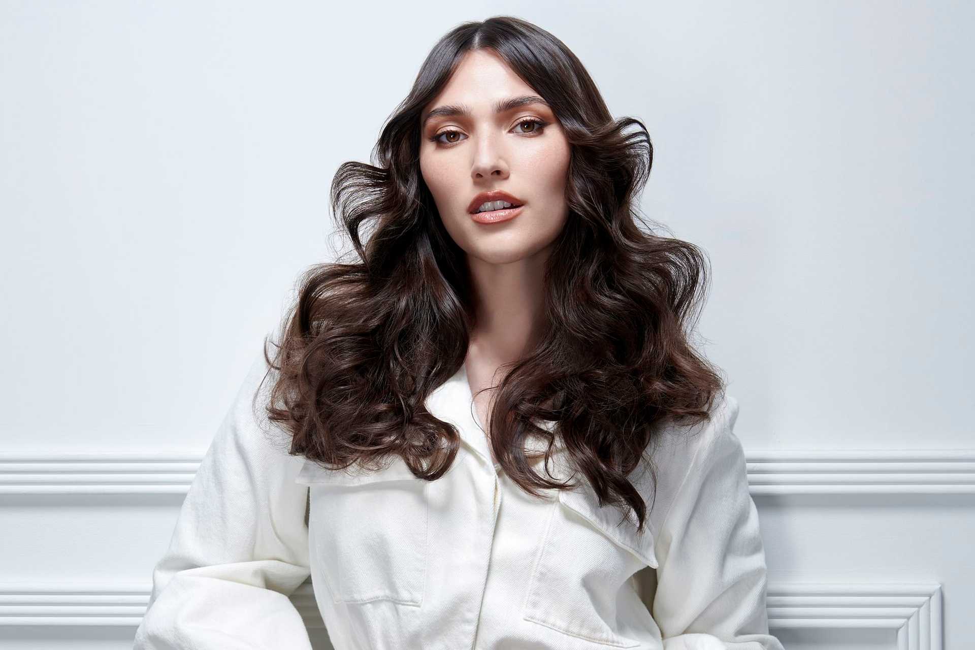 Person with long, wavy brown hair in a white outfit against a light background.