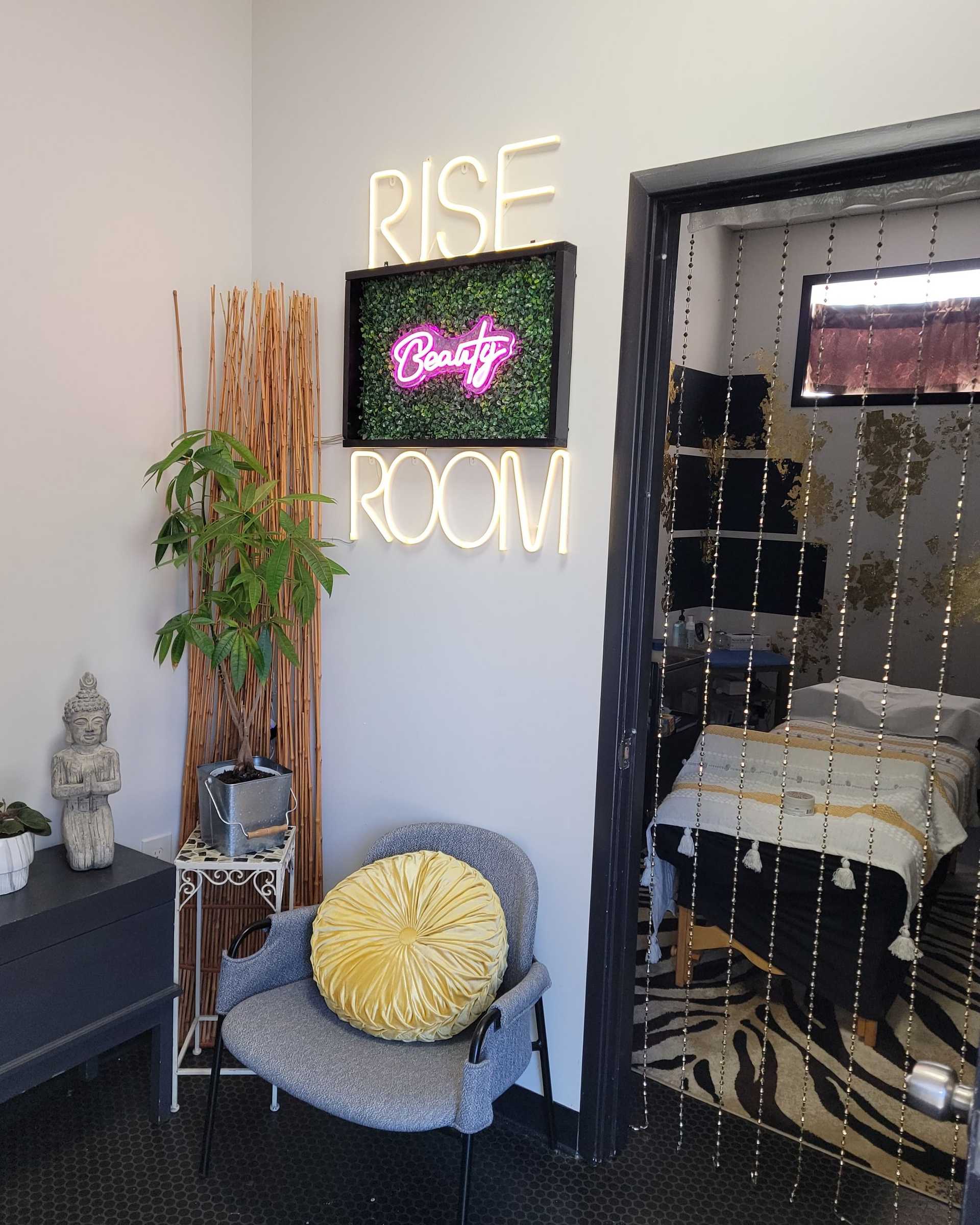 Cozy spa room with chair, plants, and neon "RISE ROOM" sign.