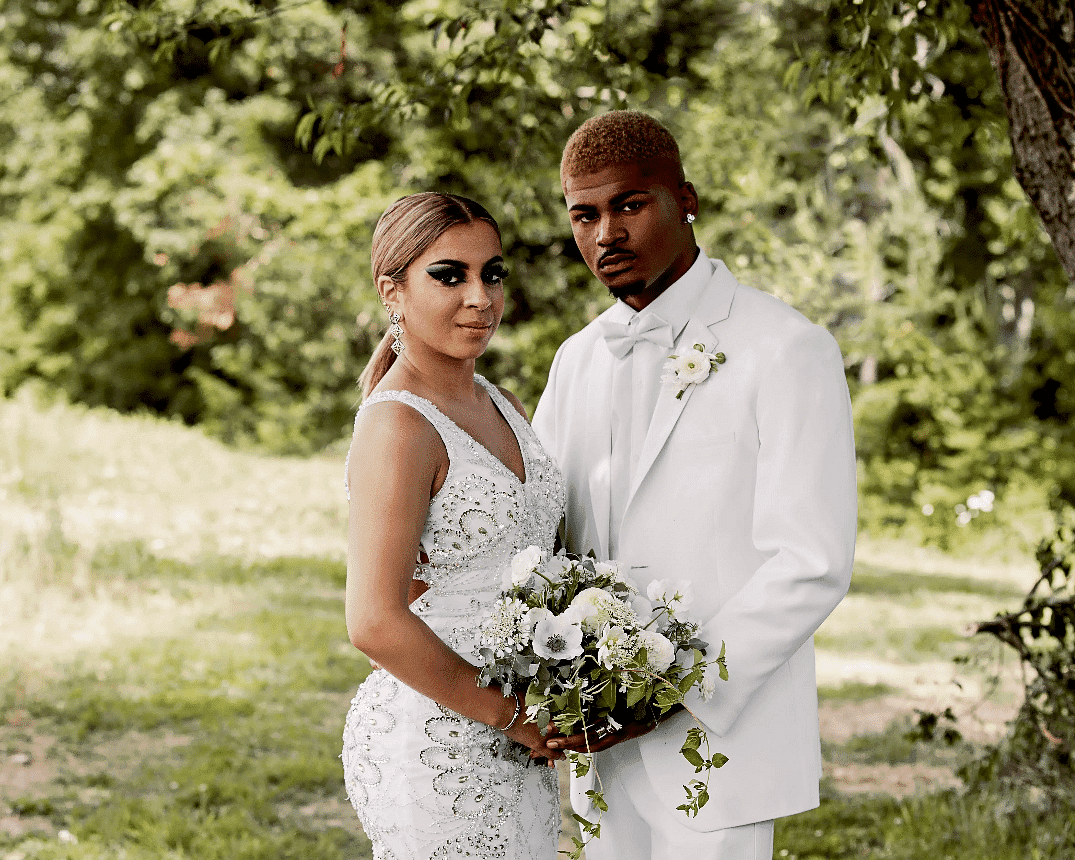 Couple in elegant white attire posing outdoors with a bouquet, surrounded by greenery.
