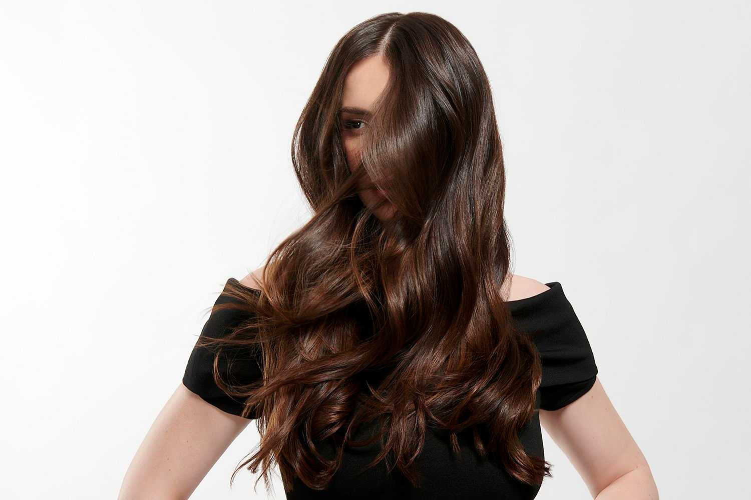 Woman with long, flowing dark hair covers her face, standing against a plain background.